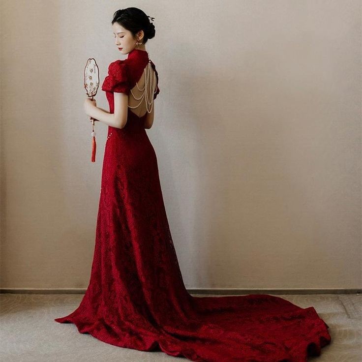 a woman in a stunning red lace gown with a train stands gracefully in a studio. the dress features a high neck, short sleeves, and a beaded back detail. she is holding a decorative hand fan and looking slightly away from the camera. the background is a simple white wall, highlighting the beauty of the dress and the woman's elegance.  the overall image conveys sophistication, glamour, and a sense of occasion.  the woman's pose and the dress's design are both elegant and captivating.  the overall impression is one of refined beauty and grace.