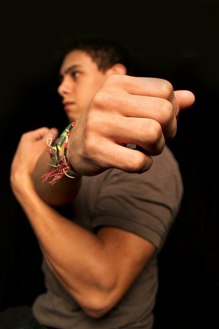 a closeup portrait of a young man with a serious expression on his face, clenching his fist. he is wearing a grey tshirt and multiple bracelets on his wrist. the image is lit from the front, creating a dramatic effect.