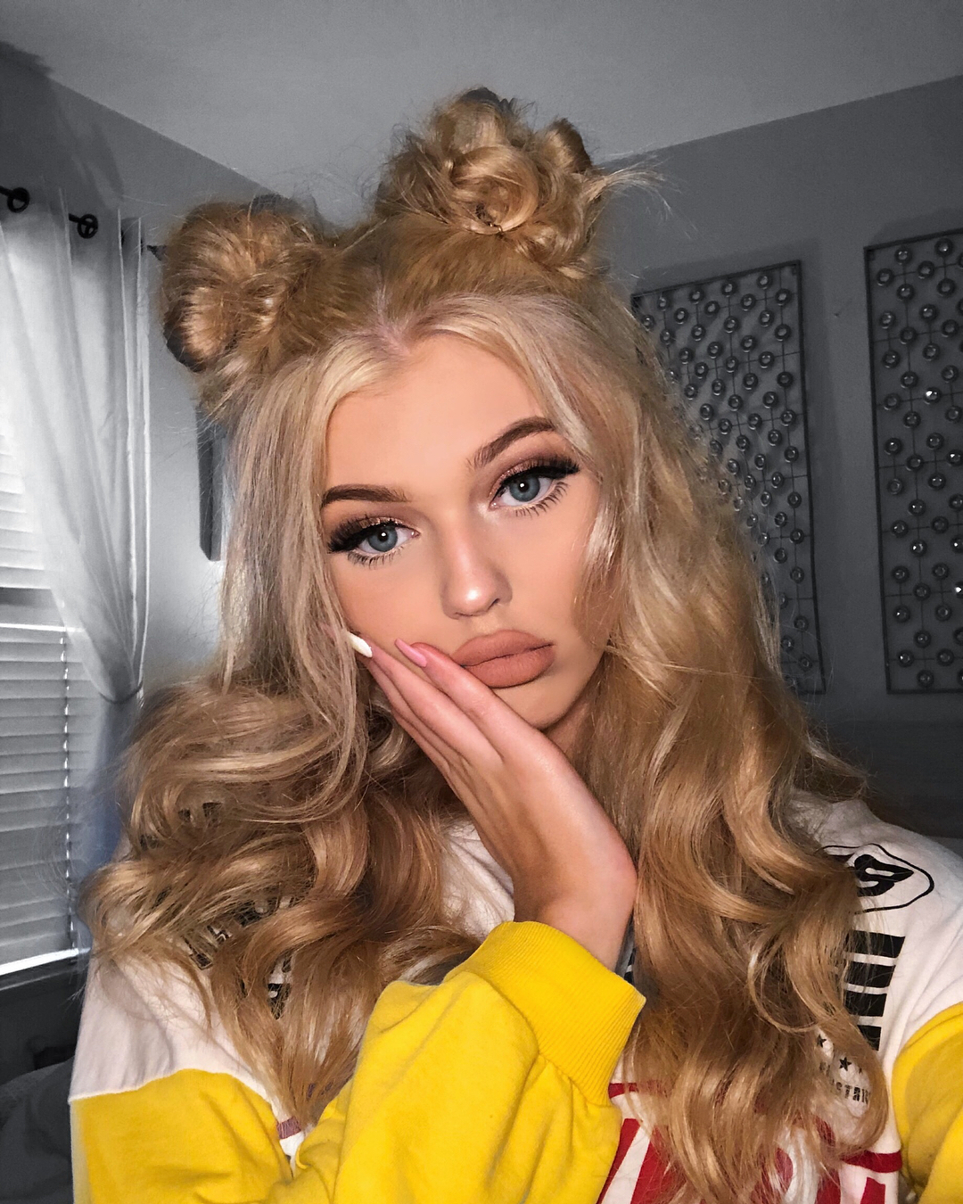 a portrait of a woman with blonde hair styled in two buns on top of her head and loose waves. she has makeup on, including false eyelashes and matte lipstick, and is wearing a yellow and white sweatshirt. her hand is on her face, and she is pouting her lips. in the background are curtains and wall decorations.