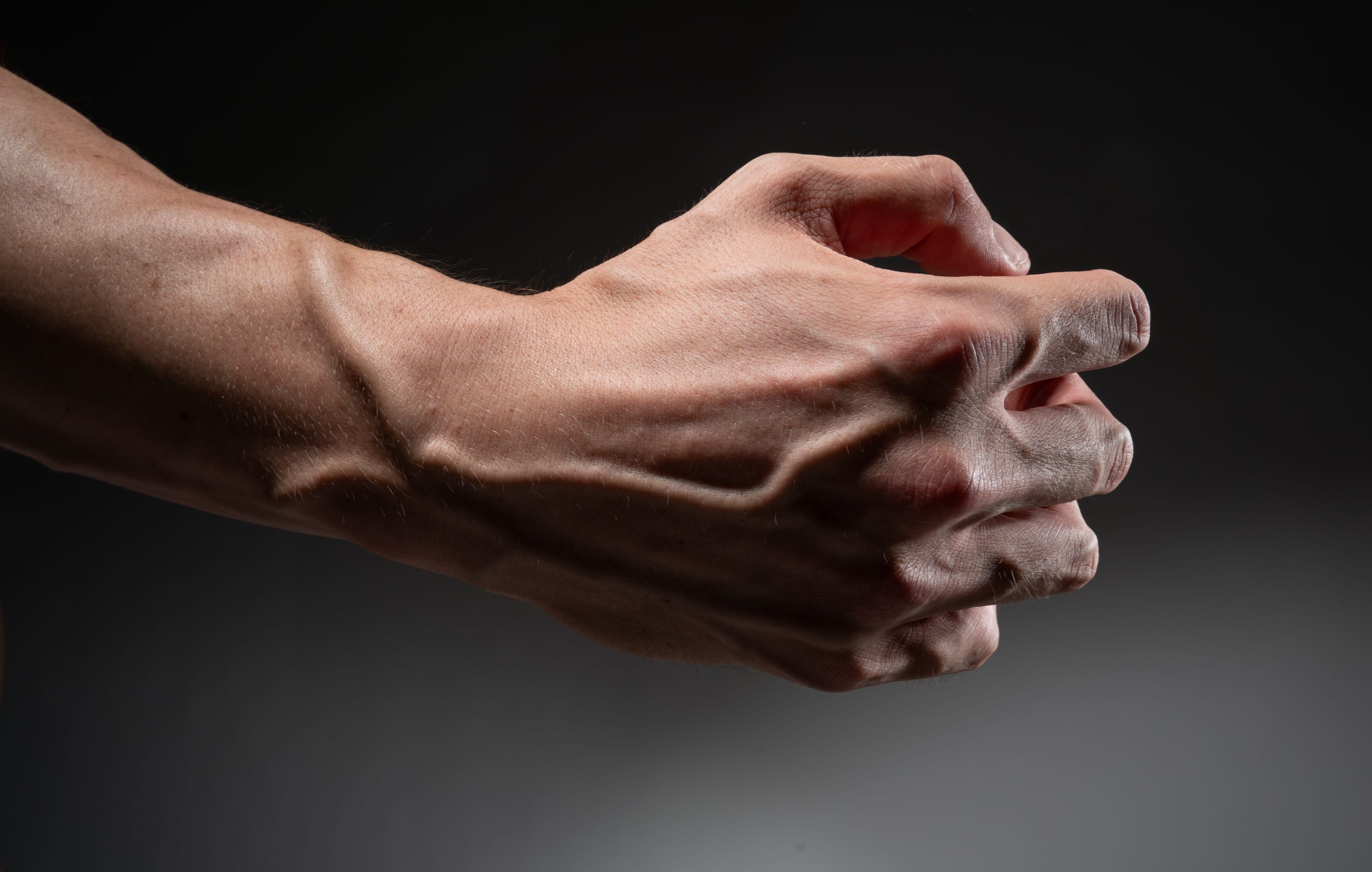 a closeup, side view of a muscular male arm and hand. the hand is partially clenched, highlighting prominent veins and vascularity. the image is dramatically lit against a dark gray background, emphasizing the definition and strength of the arm.