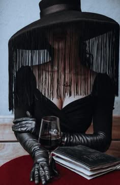 a woman dressed in black sits in a studio with a glass of wine in front of her. she is wearing a wide brimmed hat with a veil, gloves, and a dress. she has a mysterious look on her face.  the red background contrasts nicely with the black outfit, creating a dramatic look.