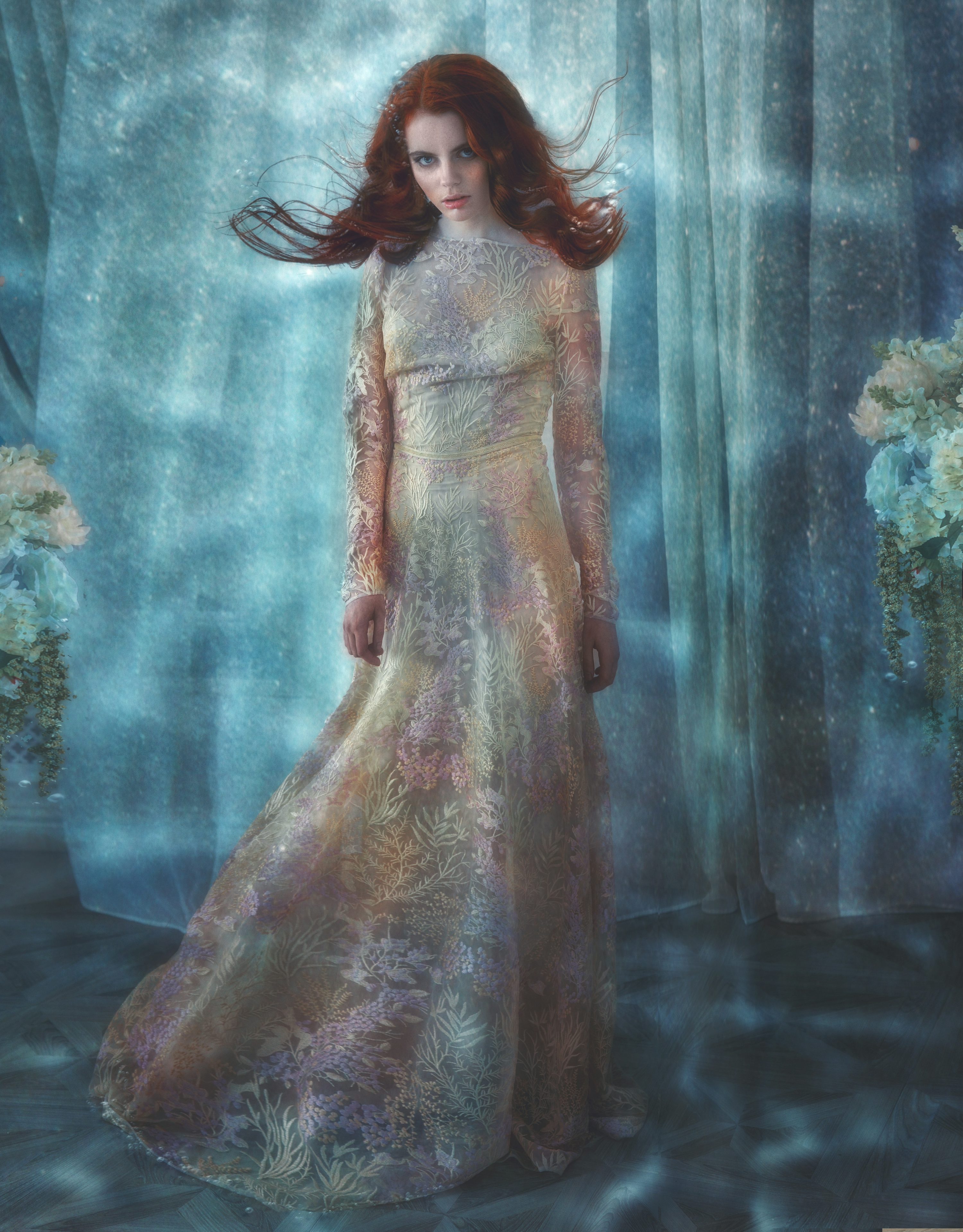 a young woman with long red hair stands in a flowing pastel floral lace gown. the setting is ethereal and mystical, with a blue and teal background suggesting a dreamy, enchanted atmosphere. the overall tone is romantic and mysterious, with a focus on the woman's graceful elegance and the beauty of her dress.