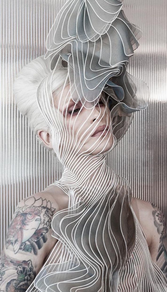 a fashion portrait of a woman wearing a futuristic and abstract outfit. she has a white and grey fabric covering her head and shoulders, creating a cool and edgy effect. she has white hair, and her face is adorned with makeup, highlighting her eyes and lips. the photo is taken in a studio setting and creates a mysterious and dramatic atmosphere.