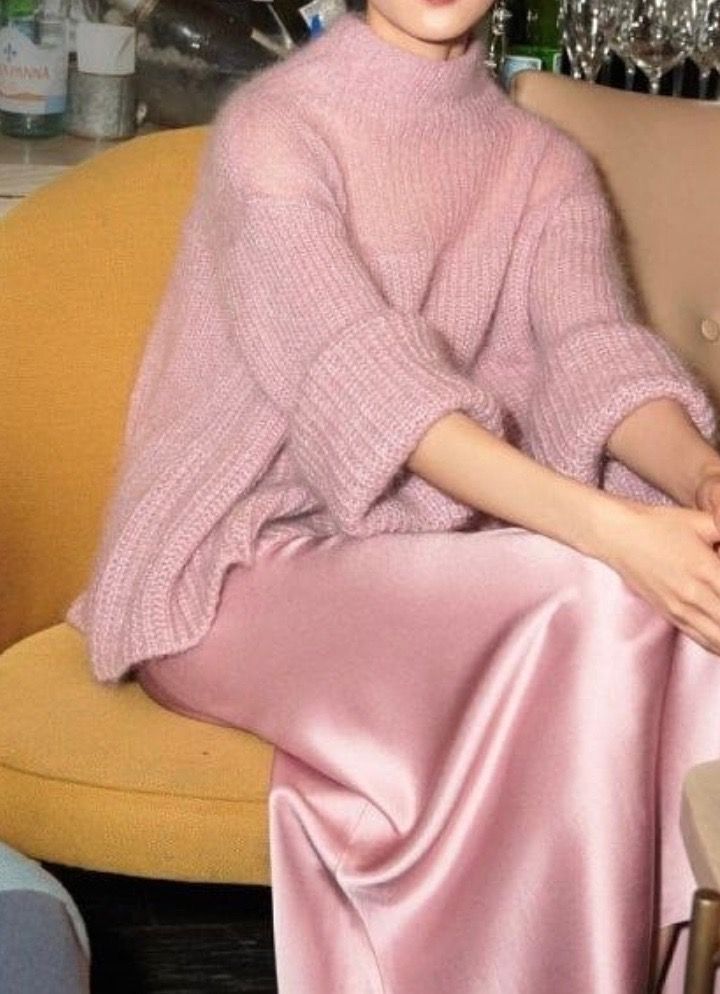 closeup view of a woman wearing a loosefitting, light pink mohair knit sweater with a high neck and threequarter sleeves.  she is also wearing a flowing, matching pale pink satin slip skirt.  she's seated on a mustardyellow chair. the setting appears to be indoors, perhaps in a restaurant or bar, with hints of glassware and bottles visible in the blurry background. the overall aesthetic is luxurious, soft, and elegant.