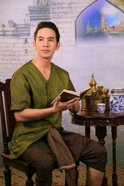 a man is sitting on a chair holding a book. he's wearing a traditional thai outfit, green shirt, and brown patterned pants. he's sitting in front of a table with teacups, vases, and other objects.  the background is a wall with traditional thai imagery.