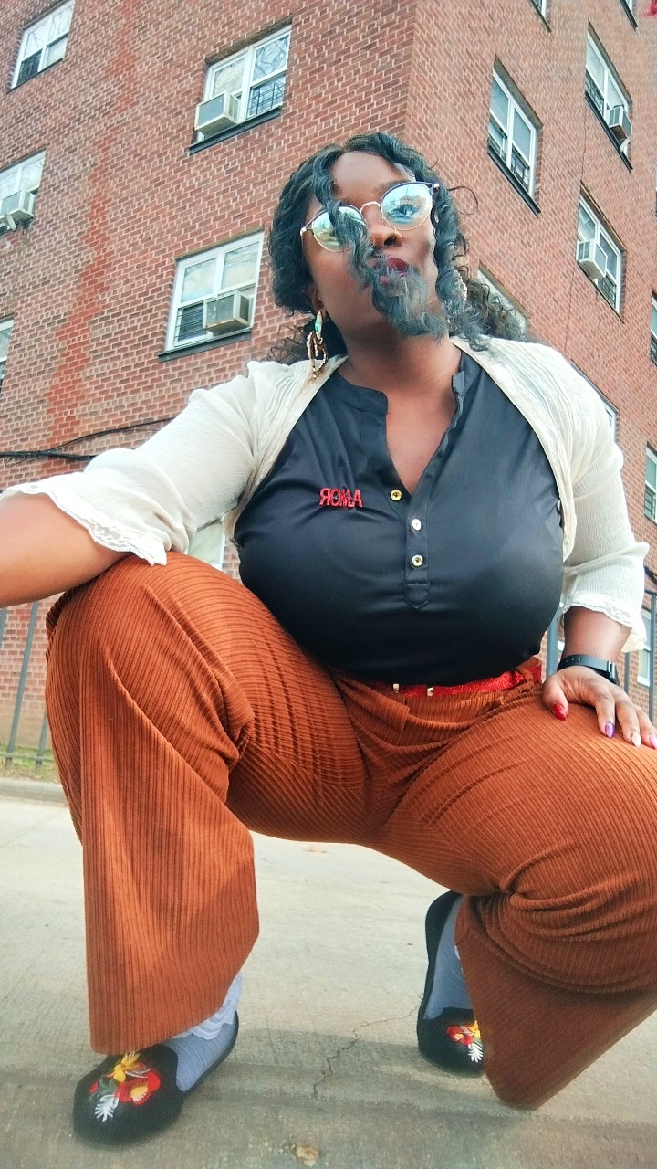 a woman in a stylish outfit poses in front of a brick building. she is wearing brown corduroy pants, a black shirt with the word "queen" embroidered on it, a white cardigan and sunglasses. her hair is styled in a big afro and she is smiling at the camera.