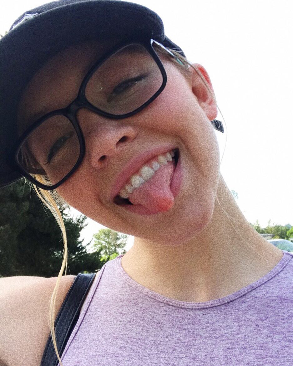 a closeup selfie of a teenage girl. she's wearing a black baseball cap, glasses, and a light purple athletic tank top. she's smiling broadly with her tongue sticking out, giving a playful expression. the setting appears to be outdoors, with some trees and part of a sky visible in the background. the image is bright and welllit, possibly taken in the daytime. the girl's blonde hair is partially visible, and her head is slightly tilted.  the overall mood is cheerful and carefree.