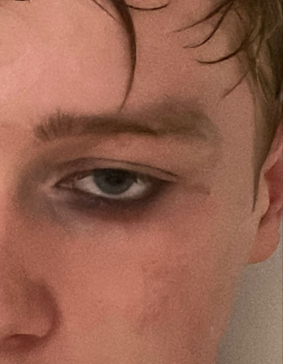 a closeup portrait of a young caucasian male's face. the subject is looking at the camera with an intense expression, showing only the eyes and brow area. the image is focused on the left eye which is heavily made up with dark eyeshadow and eyeliner.