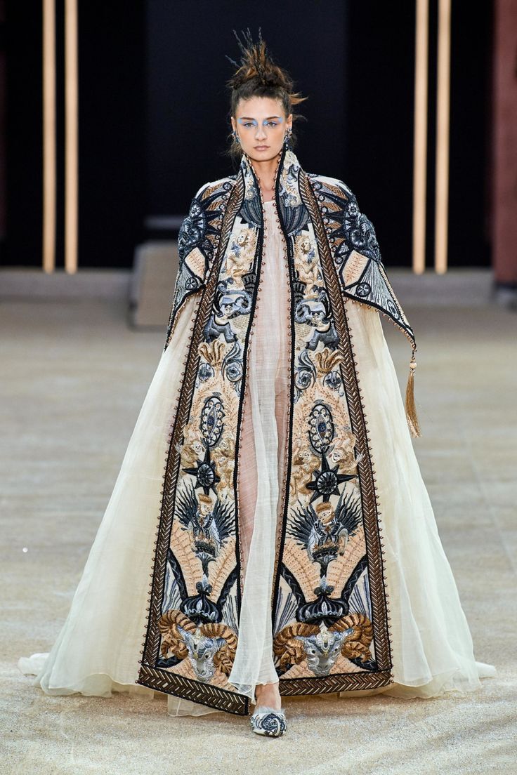 a model walks the runway in a stunning haute couture gown. the dress is a long, flowing beige creation adorned with an elaborate, intricately embroidered cape. the embroidery features a rich palette of colors and detailed designs, including gold, black, and other tones. the model's hair is styled in an updo, and her makeup is subtle yet elegant. the overall look is one of refined sophistication and high fashion. the dress is clearly a statement piece, showcasing the artistry and craftsmanship of haute couture design. the backdrop is a neutraltoned runway, allowing the garment to take center stage.