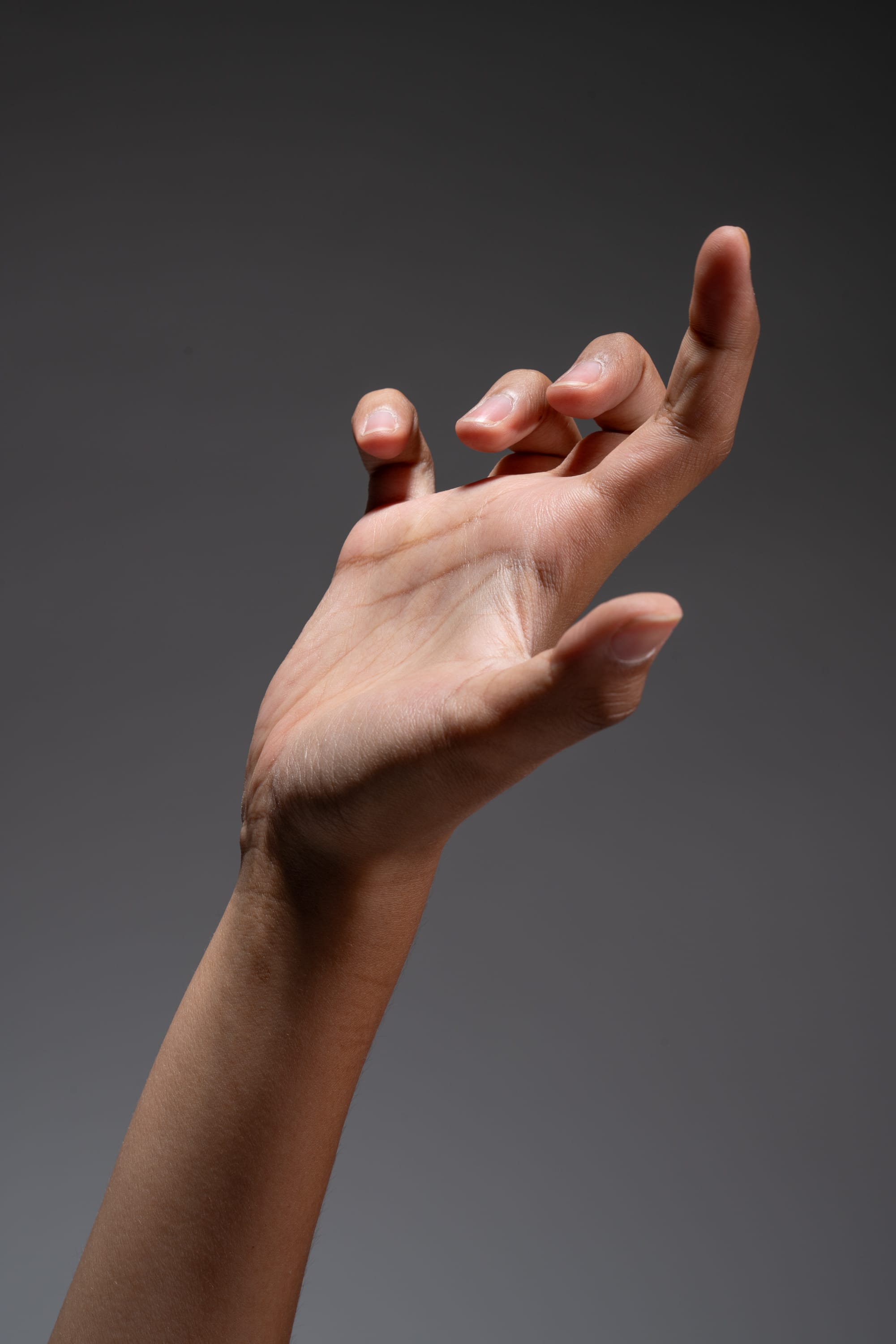a closeup image of a hand reaching upward against a gray background. the hand is lit with a spotlight, creating a sense of tension and drama. the fingers are curled slightly, as if the hand is grasping at something.