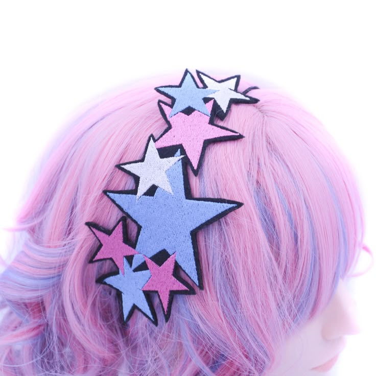 a closeup image shows a pink and blue wig with a hair accessory featuring a cluster of embroidered stars in various sizes and pastel colors (pink, blue, white) outlined in black. the stars are positioned on the top of the wig, close to the forehead.