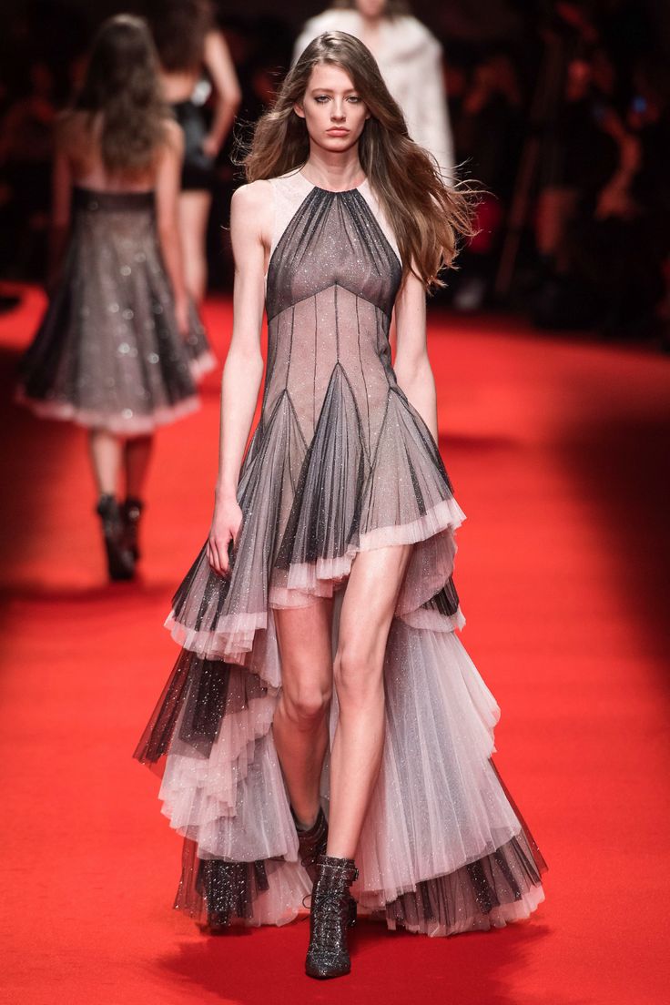 a model walks the runway in a stunning highfashion dress. the dress is a highlow design, with a layered tulle skirt in shades of black and pink. the bodice is fitted and sheer, and the overall effect is both elegant and glamorous. the model's dark hair and makeup complement the dress, and her confident stride adds to the overall impact. the background is a red carpet, typical of a highfashion show. the picture shows a woman with long hair wearing a layered, highlow tulle dress walking on a red runway. the dress is mostly black and light pink tulle, with a hint of glitter. she is wearing black ankle boots.