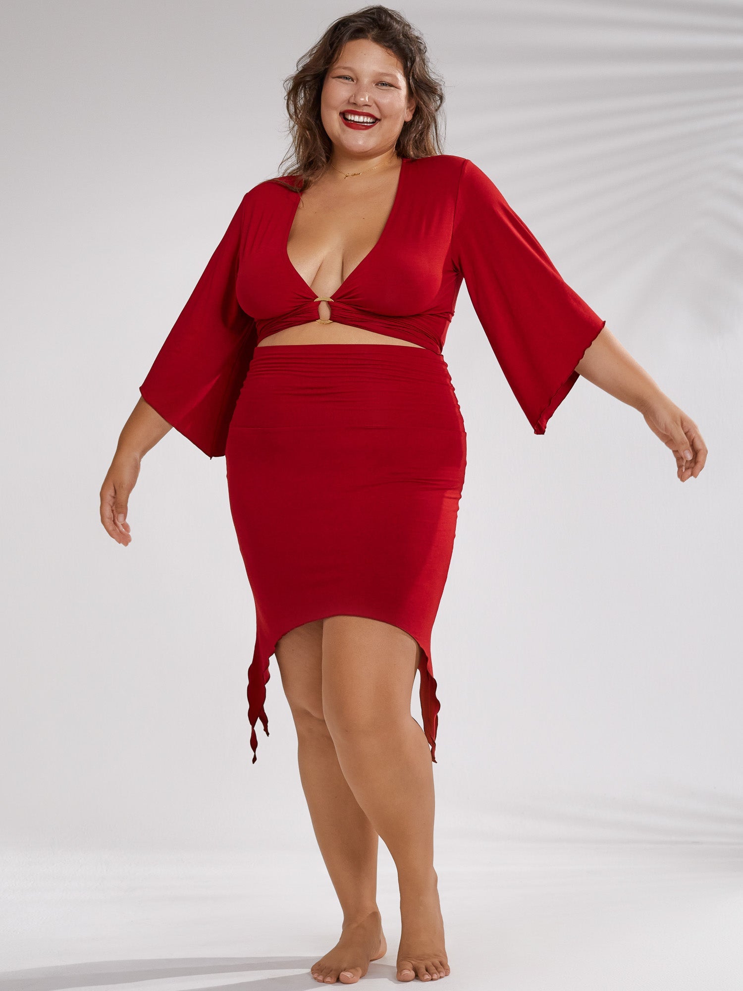 a full shot of a plussize model wearing a stylish red dress. the dress features bell sleeves, a vneckline with a cutout detail, and an asymmetrical hemline that is longer in the back and shorter in the front. the model is smiling and standing confidently against a white background. the overall style is elegant and sophisticated, highlighting the beauty and confidence of plussize women.