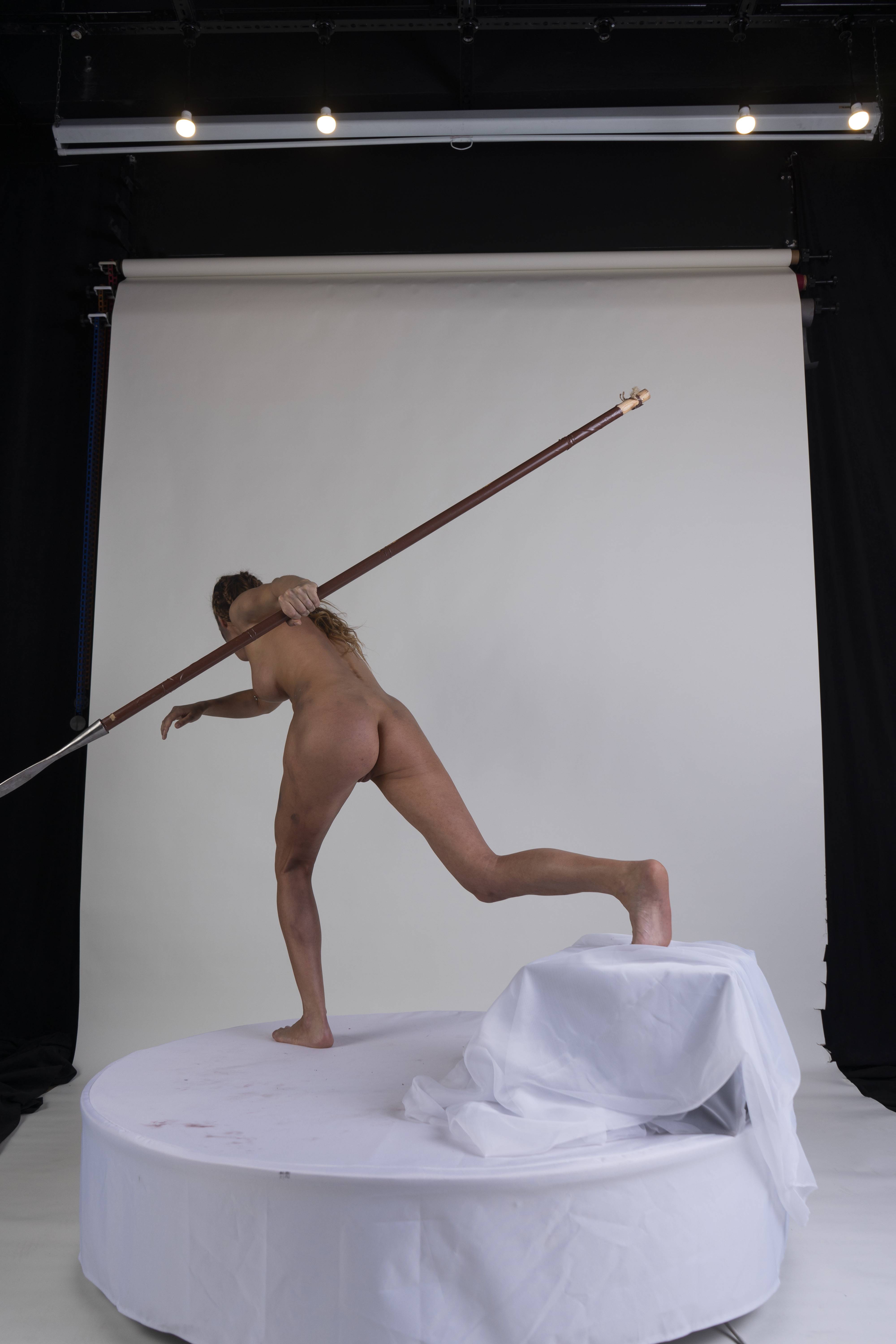 a nude woman stands on a platform in a studio setting, holding a spear and posing in a dynamic, athletic stance. the shot is taken from the back, showing her full body.