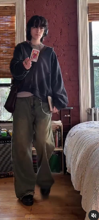 a young woman with long black hair and bangs is standing in a bedroom.  she is wearing a gray sweatshirt and jeans.  she is holding a card and a book in her hands. she is looking to the side.  there is a bed in the background.