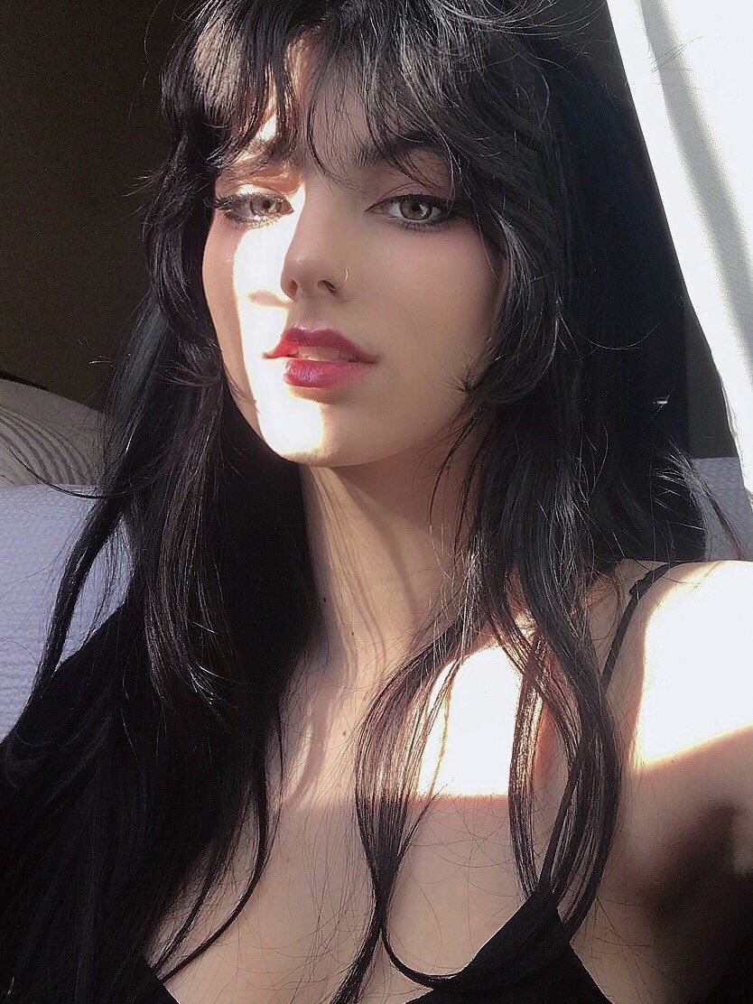 the image is a closeup portrait of a young woman with long, dark hair styled with curtain bangs that frame her face.  sunlight streams in from the side, casting shadows and highlighting her features. she has green eyes, subtle makeup, and a small nose ring. she's wearing a simple black top that reveals her shoulders and neck. the overall aesthetic is soft, dreamy, and slightly mysterious. the lighting and styling create a mood of elegance and beauty.