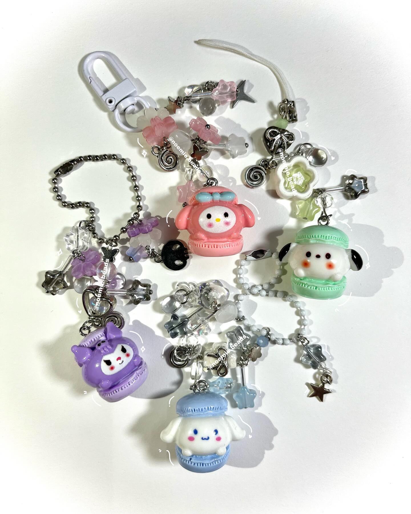 a closeup shot showcasing five handmade keychains featuring kawaiistyle sanrio characters transformed into "macaron" style charms. each charm is attached to chains and various beads in pastel colors and various shapes like stars and butterflies.