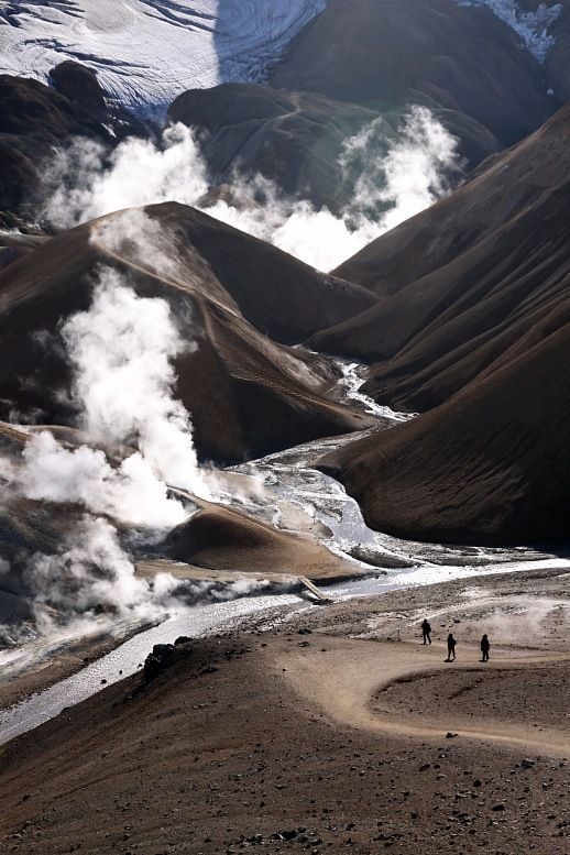 the image shows a landscape with mountains, a stream, and steam rising from the ground in a geothermal area. the scene includes brown hills, a snowcapped mountain range in the background, and three hikers walking on a path. it appears to be taken in iceland, possibly in landmannalaugar, with a focus on the volcanic landscape and geothermal activity.
