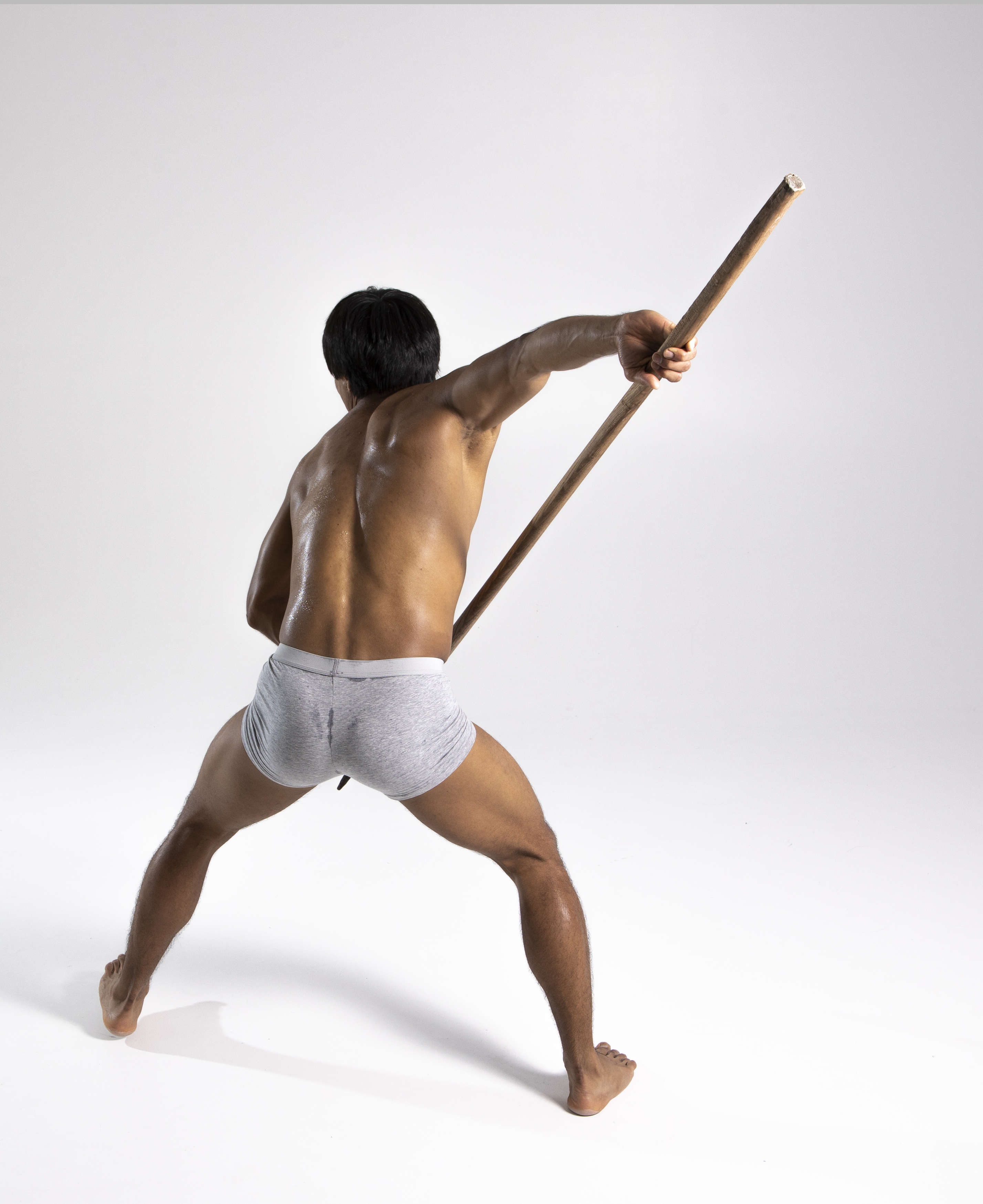 a shirtless man is seen from the back, posing in a warrior stance while holding a wooden staff. he is wearing gray boxer shorts and is barefoot. the background is a plain white studio backdrop.