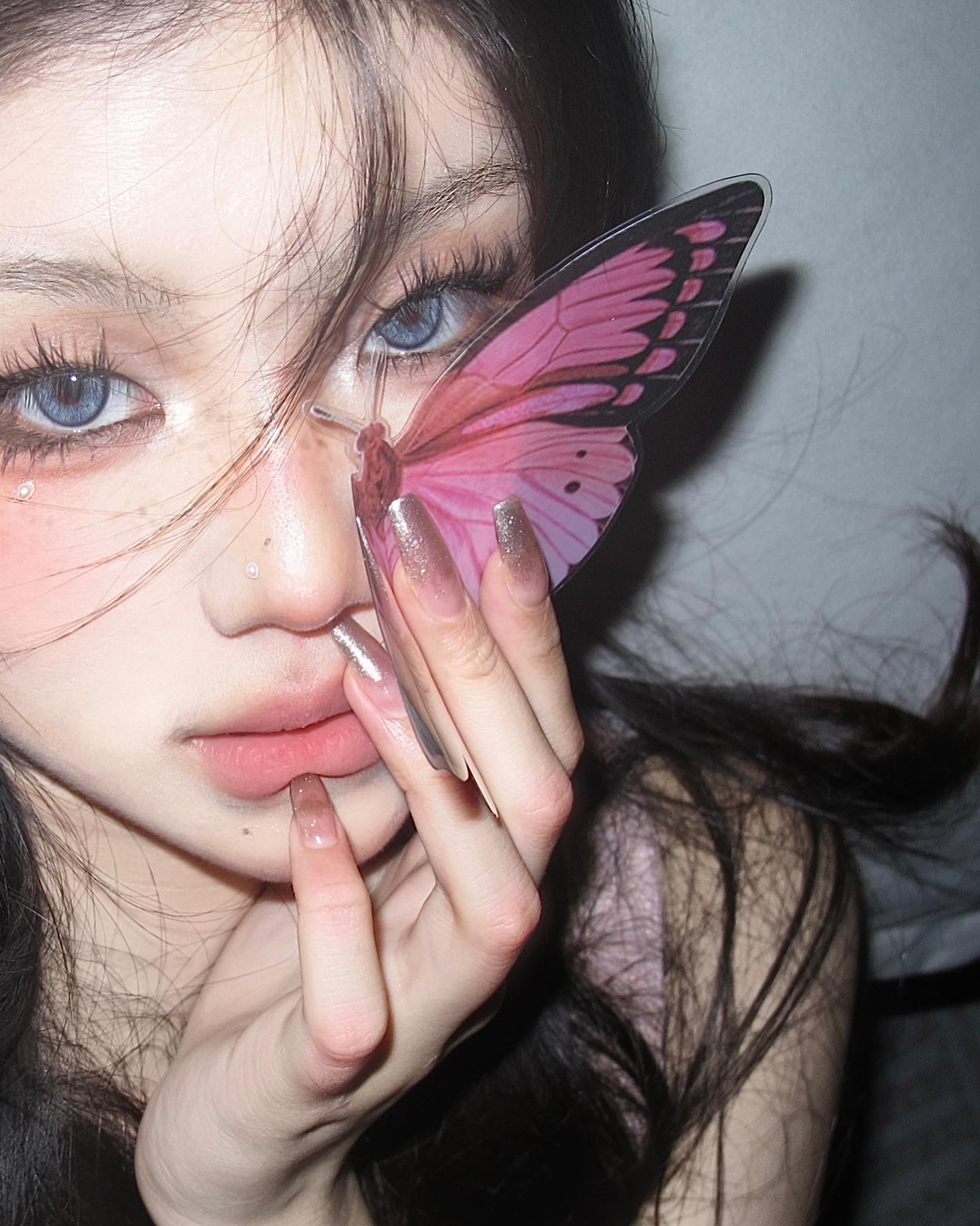a closeup selfie of a young woman with a butterfly graphic partially obscuring her face. she has dramatic eye makeup with blue contacts, pink blush, and glossy lips. her nails are long and feature a glittery pink ombre design. her expression is serious and alluring.