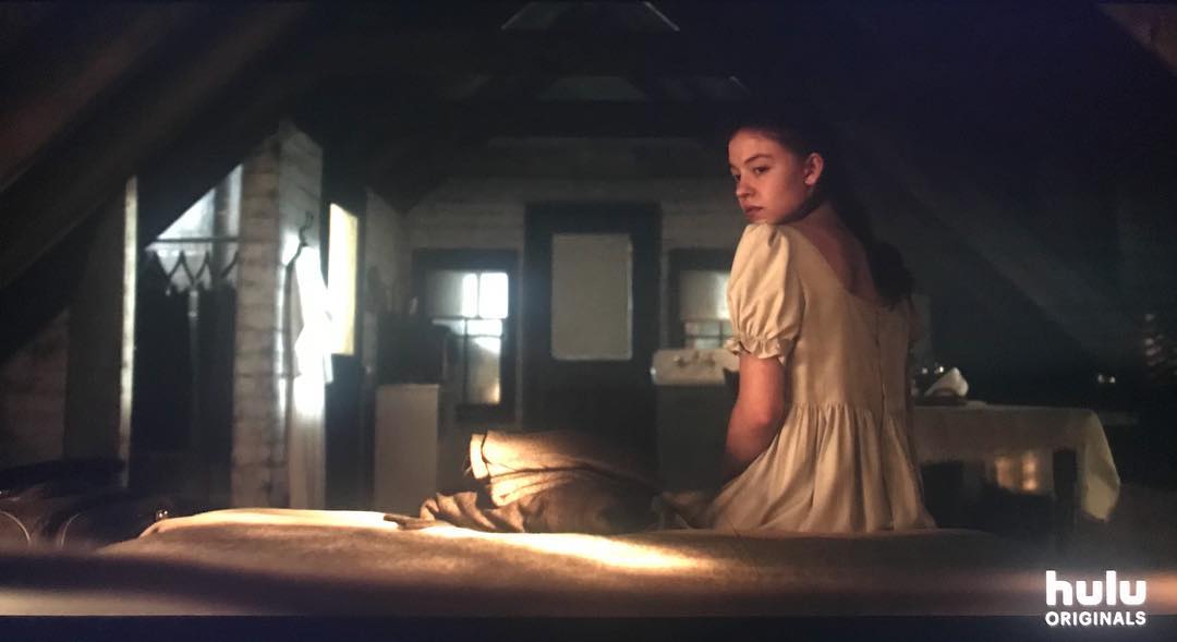 a still image from a hulu originals show. a young woman in a lightbeige dress sits on a bed in a dimly lit attic bedroom. her back is to the camera, and she is looking away. the room is old and slightly rundown, with antique furniture and a general sense of mystery and suspense. the lighting is low, creating shadows and an atmospheric mood.  the hulu originals logo is visible in the bottom right corner.