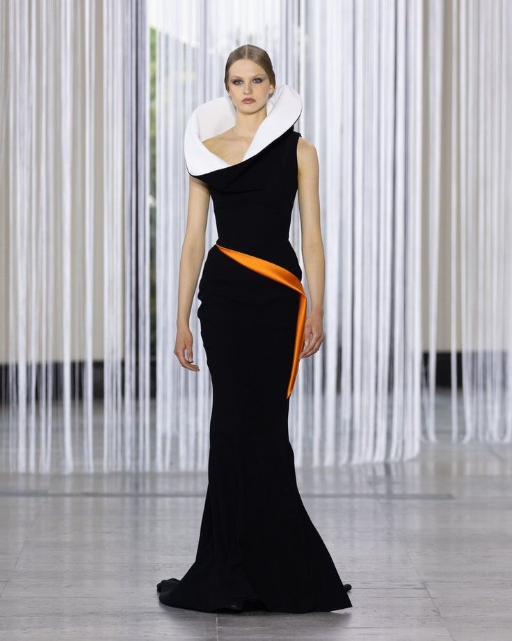 a model walks down a runway in a black dress with a dramatic white collar and an orange sash. she is wearing her hair pulled back from her face. the backdrop is a white wall with vertical lines.