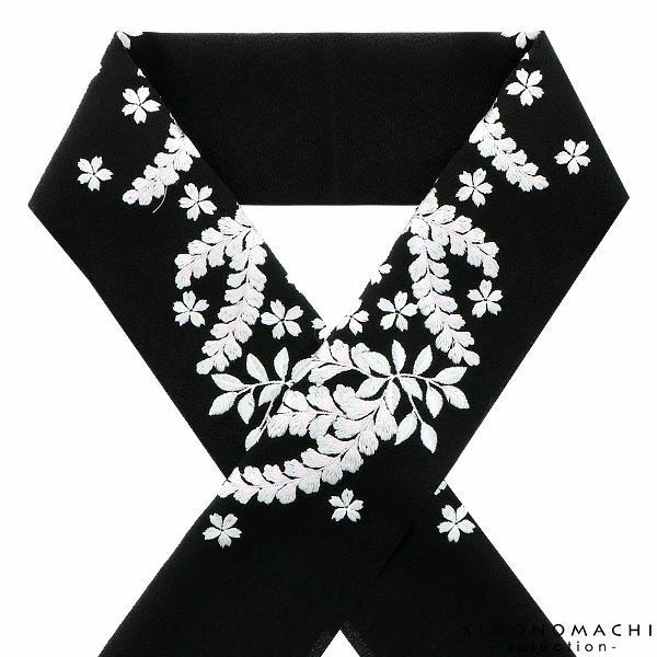 a black obi, or kimono belt, is displayed with an embroidered white floral pattern against a white background. the obi is folded in a v shape, with the floral design concentrated towards the center and edges of the visible portions. the lower right corner has a small logo or watermark.