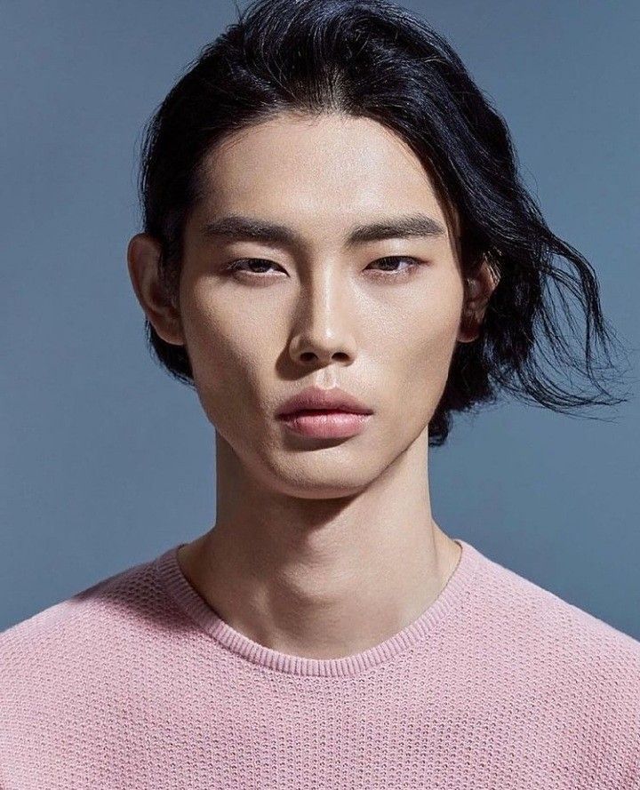 a portrait of an asian man with long, black hair wearing a pink sweater. he is facing the camera in a studio setting. he has light skin and a neutral expression.