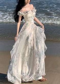 a woman in a white floral dress stands on a beach with the ocean behind her. she is smiling and looking happy. the dress is flowing in the wind and her hair is blowing in the breeze. the sky is blue and the sun is setting in the background. the woman looks carefree and romantic.