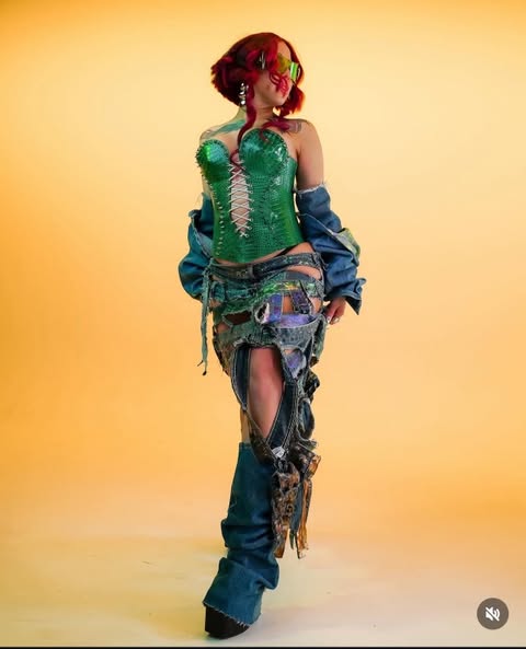 a woman stands in a fullbody shot against a yellow background. she is wearing a green snakeskin corset, a denim jacket with the sleeves pushed up, a ripped denim skirt, and denim boots. her hair is red, and she is wearing sunglasses and earrings. she is looking away from the camera.