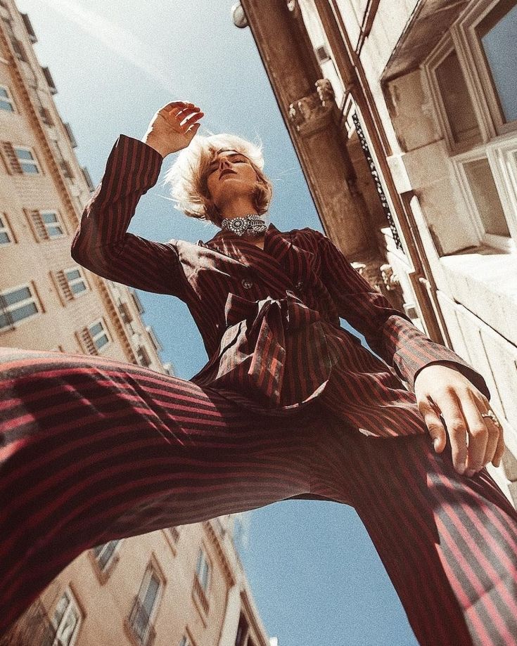 a woman wearing a striped suit, stands on a city street, looking up at the sky. she is in the middle of a wide shot, with buildings on either side of her. the perspective is low, making the buildings look tall and imposing. the woman's face is obscured by her hair. she is wearing a ring on her finger.  the image has a retro feel to it.