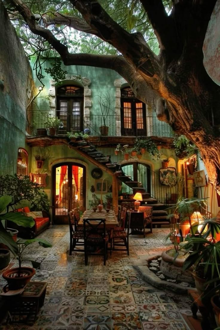 this is a view of a lush, rustic courtyard, likely in a traditional setting. a large tree dominates the space, casting shadows over the tiled floor. the building features a balcony with potted plants, stone accents around the windows, and wooden stairs leading to an upper level. a dining table with chairs is set up in the courtyard, illuminated by a lamp, and various plants contribute to the cozy, inviting atmosphere.