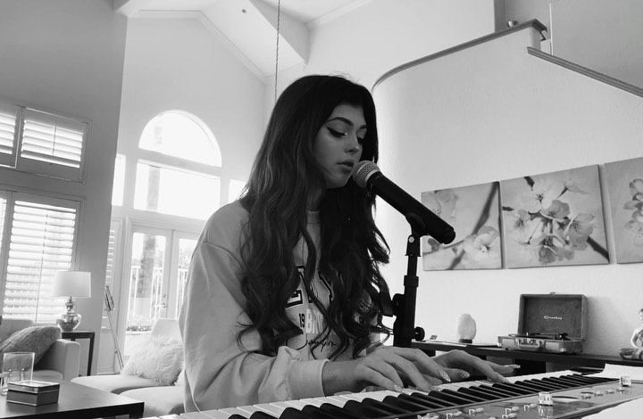 a black and white image shows a young woman playing a piano and singing into a microphone. she has long dark hair and is wearing a sweatshirt. behind her are some paintings, a record player, and a staircase. there is a couch and window in the background.