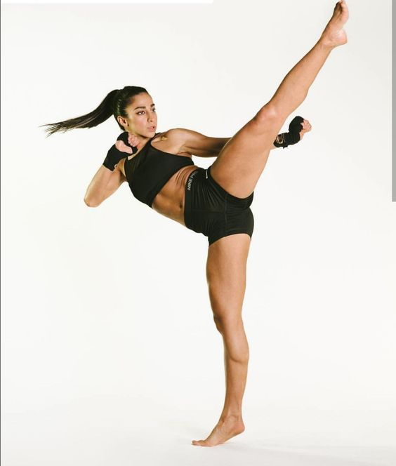 a woman in athletic wear executes a powerful high kick against a white background. she is wearing black shorts and a black sports bra, her long dark hair is tied back in a ponytail, and her fists are clenched. the image captures her strength, flexibility, and athleticism.