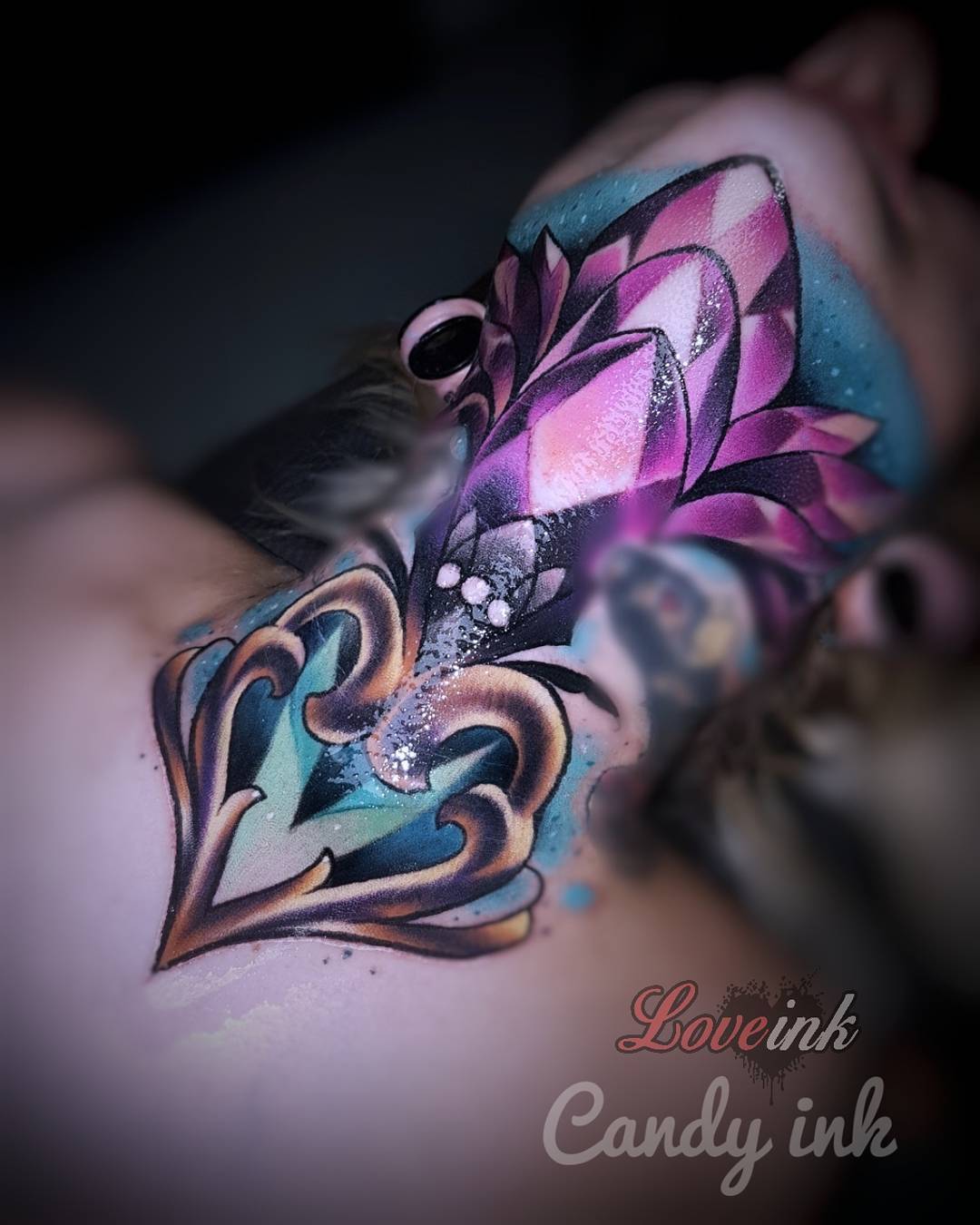 a closeup image showcasing a pink and blue jeweltoned tattoo design drawn on a woman's neck. the tattoo design features a heartshaped base with an ornate, diamondlike jewel on top.  there is a watermark in the bottom right corner that reads "loveink candy ink".