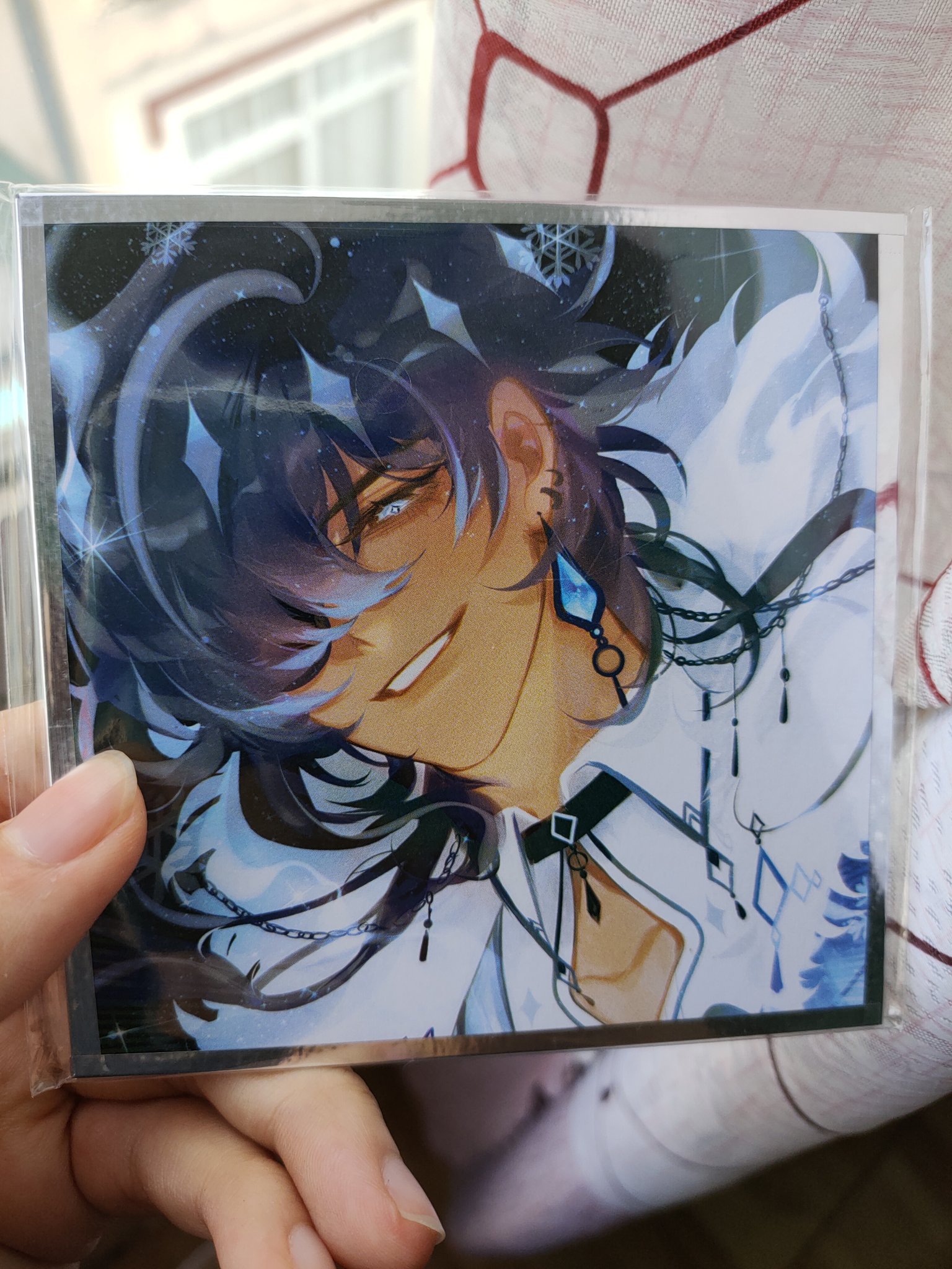 the image shows a person's hand holding a clear plastic sleeve containing a printed piece of fanart. the artwork depicts a stylized animestyle male character with long, dark blue hair. he has light skin, and is wearing a lightcolored, possibly white, shirt or coat with intricate detailing,  and multiple necklaces and earrings with dark and light blue accents. the background is dark with hints of blue and white, suggesting a winter or snowy setting with snowflakes. the art style is clean and detailed, with a focus on the character's expression and clothing. the image is a closeup, emphasizing the quality of the print and the artwork's details.  the hand holding the print is partially visible, as is a portion of a lightcolored garment beneath it.
