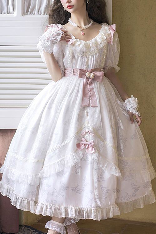 a young woman models a white lolita fashion dress. the dress is floor length and has a layered, ruffled skirt with lace trim. the bodice features a sweetheart neckline, puffed sleeves, and a pink ribbon tied at the waist.
