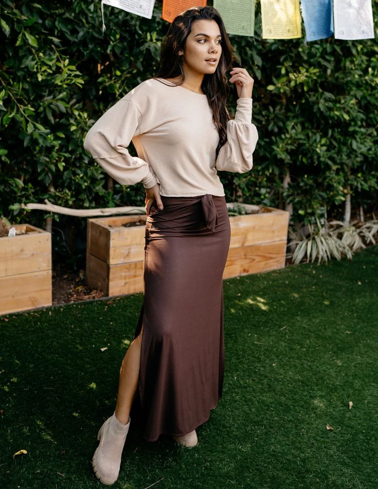 a woman in a beige top and a brown side slit skirt is standing on a lawn in a garden. she is wearing white ankle boots. the image is taken during the day with natural lighting. the perspective is at eye level.