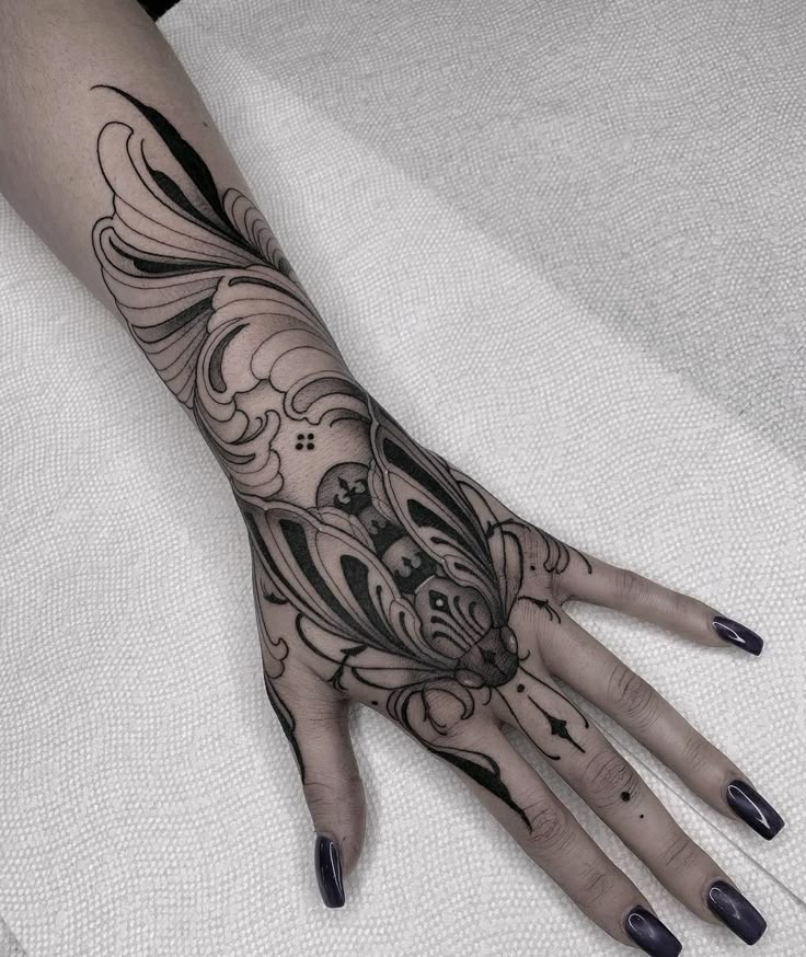 a closeup shot of a person's hand and part of their forearm, featuring an elaborate black ink tattoo that covers the majority of the hand and extends up the arm. the tattoo's design includes a mothlike figure with intricate details, ornate patterns, dots, lines, and shading. the person's fingernails are painted with a dark nail polish. the hand rests on a white, slightly textured surface, possibly a cloth or paper background.