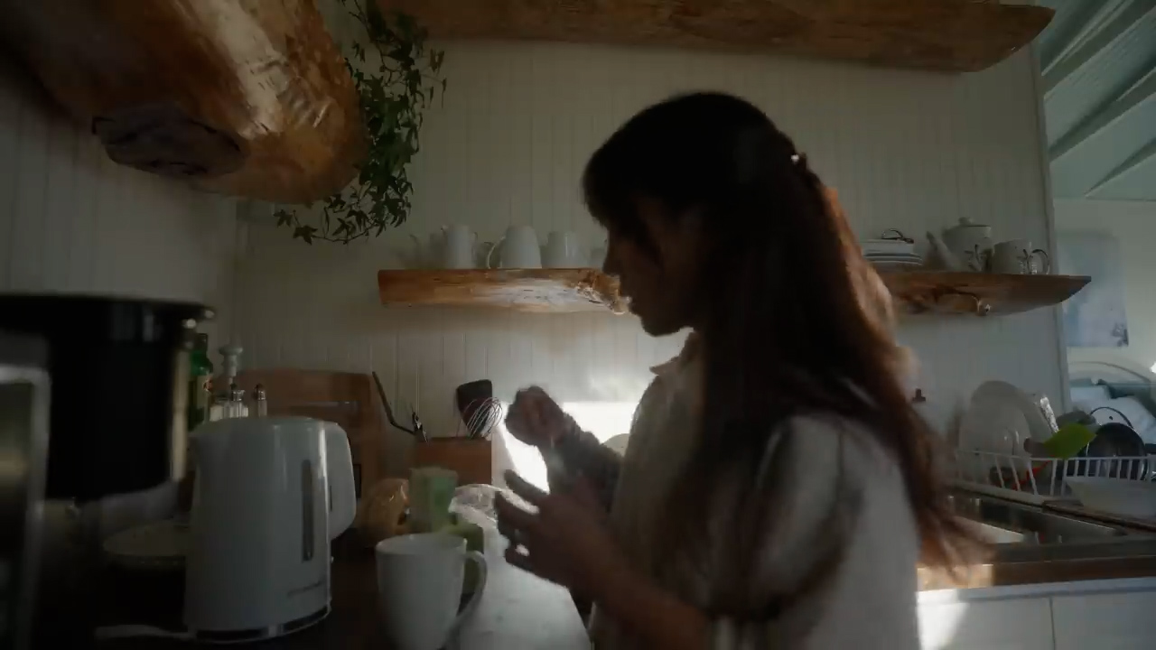 a woman with long dark hair is standing in a brightly lit kitchen. she's wearing a white shirt and is making herself a cup of coffee. the kitchen has wooden accents and white walls. there is a coffee maker, a kettle, and various mugs on the counter.