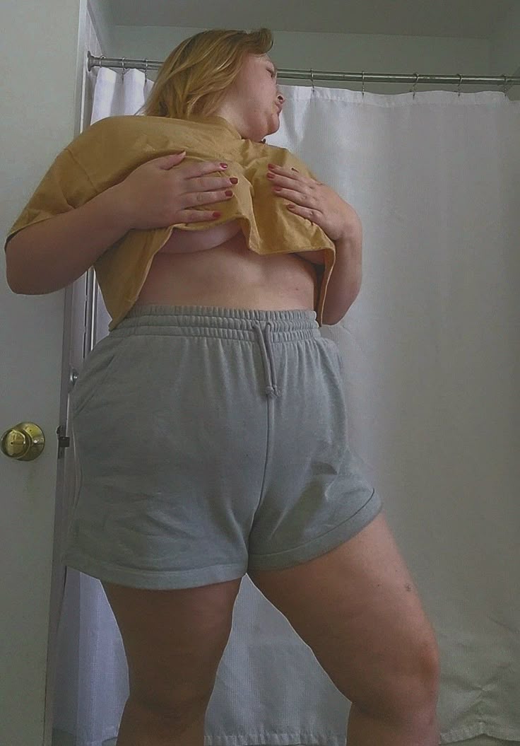 a woman in a crop top and shorts is standing indoors. her hands are covering her chest. the background is a shower curtain and door.