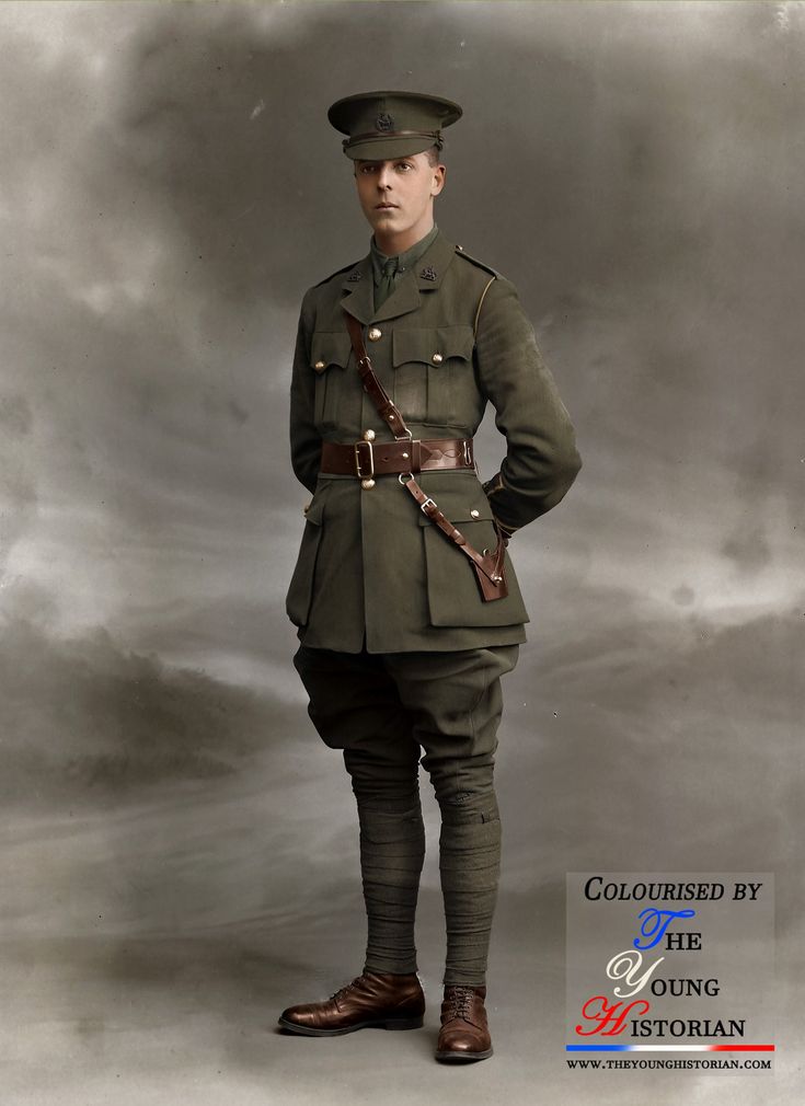 a full shot of a man in a military uniform from what looks to be world war i. he is wearing a hat, a uniform jacket, a belt, riding breeches, leggings, and boots. he is standing with his hands clasped behind his back. the background is a cloudy sky. the image is colorized. there is a logo on the lower right.