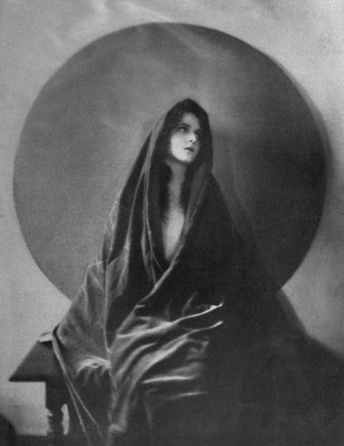 this is a vintage portrait of a woman in a dark shawl. she is posed in a dramatic fashion, and the lighting is high contrast, creating a sense of mystery. the woman's face is partially hidden by the shawl, which adds to the air of intrigue. the image is likely from the 1920s and has an art deco feel. the photograph is black and white and  has a grainy texture, further adding to its vintage charm.