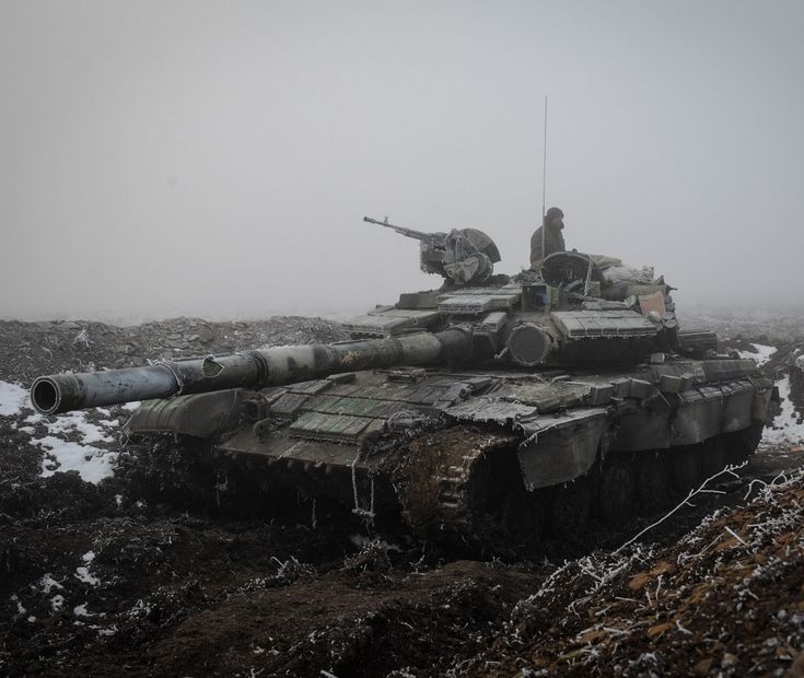 a closeup view of a russian t90m proryv main battle tank, partially obscured by fog and covered in mud and some frost. a soldier is visible inside the tank's turret.  the tank appears to be in a field of dark earth and patches of snow, suggesting a cold, possibly wintery battlefield environment. the tank shows signs of wear and tear. the overall mood is one of seriousness and military conflict.