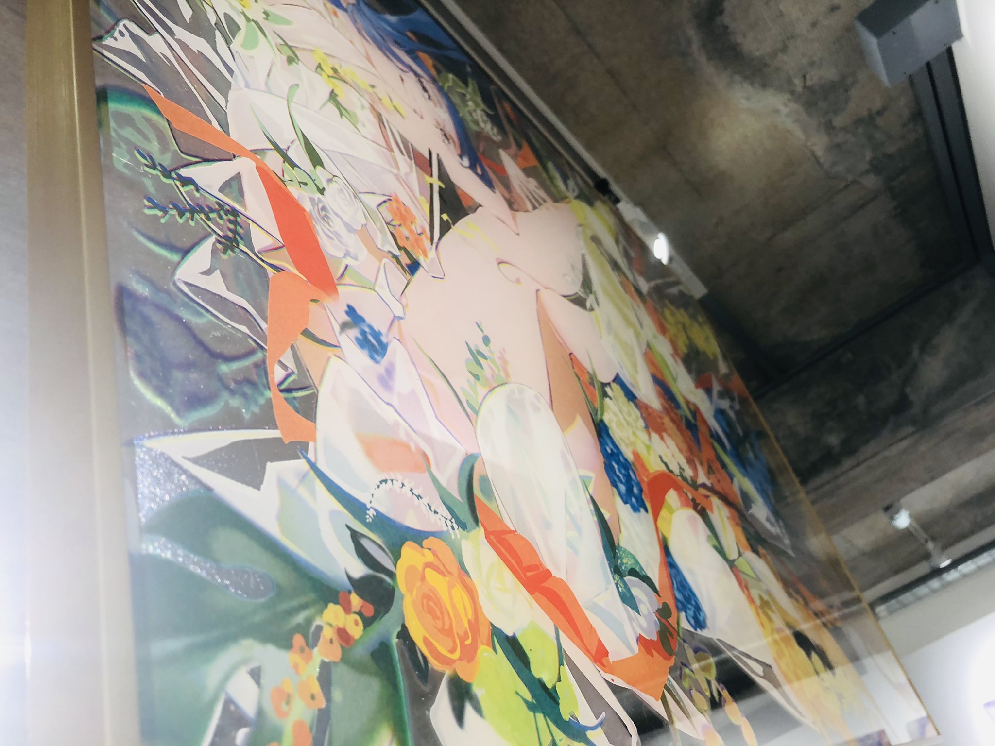the image shows a large, colorful, abstract painting in a gold frame. the painting is mounted on a wall with a concrete ceiling. the painting appears to depict a figure partially obscured by abstract shapes and flowers in various colors, including white, red, blue, pink, yellow, and green. the painting style is modern and somewhat fragmented, with overlapping and translucent elements. the image is taken from a low angle, looking up at the artwork, which emphasizes its size and presence in the gallery space. the lighting is bright, suggesting natural light from a source like a skylight or large window.