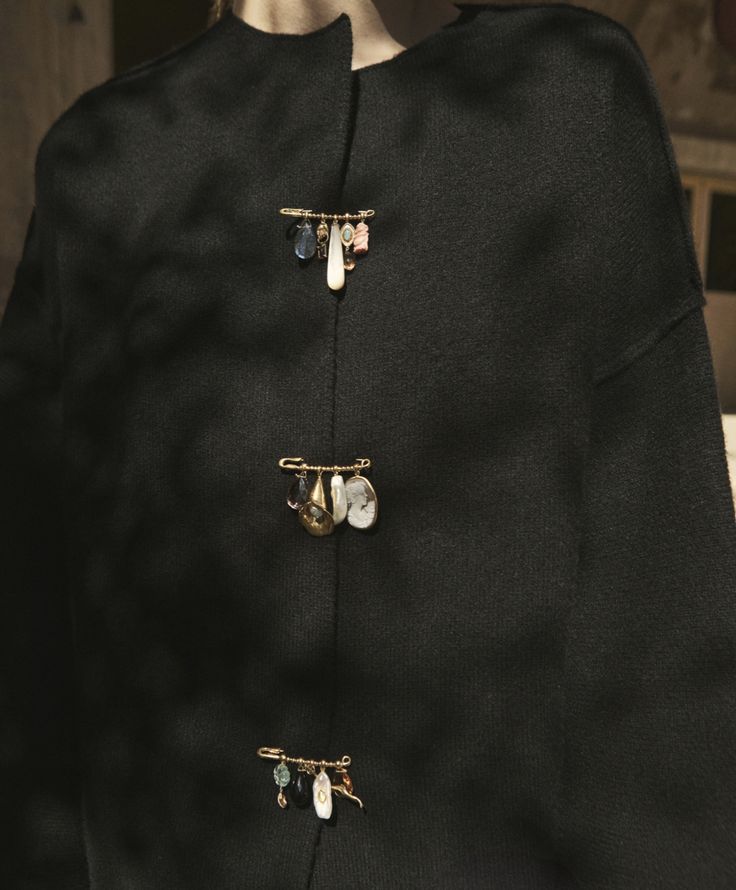 a closeup shot showcases a black cardigan fastened with three decorative goldcolored safety pins. each pin is adorned with various charms, including beads and pearls, adding a personalized and artistic touch to the garment.