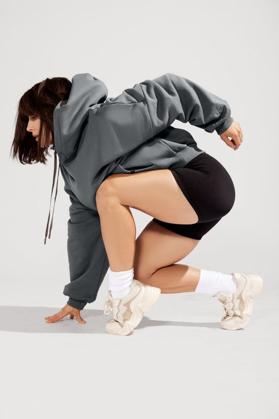a young woman wearing a gray oversized hoodie, black athletic shorts, and white chunky sneakers is dynamically posed against a light gray background. she is crouched low with one leg extended and her hand touching the floor, as if about to spring into action. the studio lighting is bright and emphasizes the contours of her athletic build and the casual yet stylish fit of her activewear.