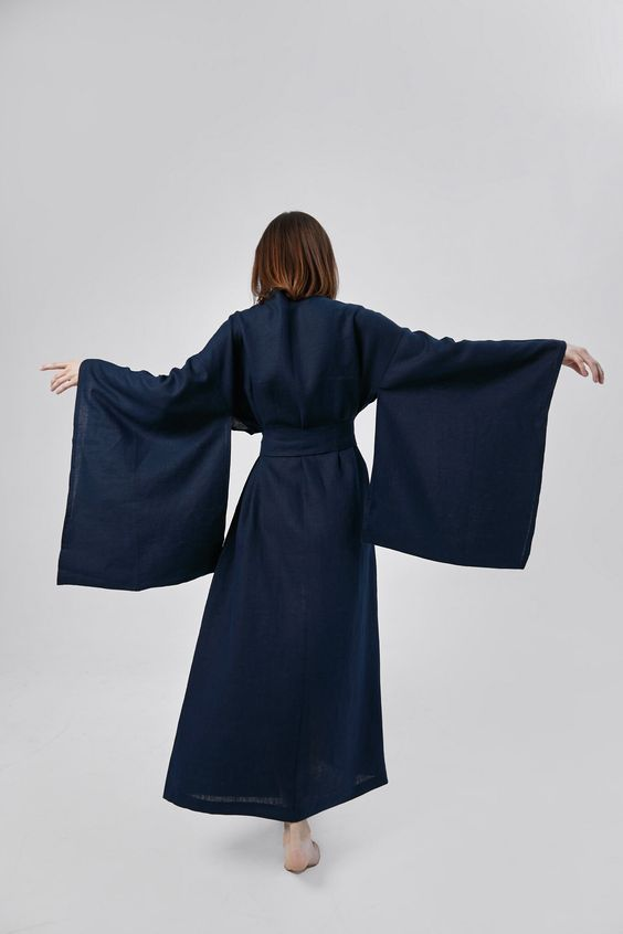 a woman with shoulderlength brown hair stands with her back to the camera, showcasing a long, flowing navy blue kimono dress. the dress features wide, dramatic sleeves, a tie waist, and falls to her ankles. it's made of a natural fabric that drapes elegantly.  the studio setting provides a clean, white background that allows the rich color and details of the dress to stand out.