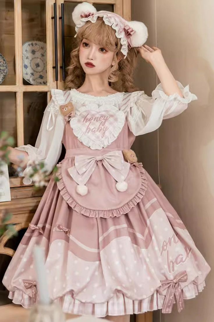 a young woman poses in a pink and white lolita fashion dress. the dress is adorned with lace, bows, and teddy bear details. she is also wearing a matching headband with bear ears. the background is a wooden cabinet and the photo is lit with studio lighting.