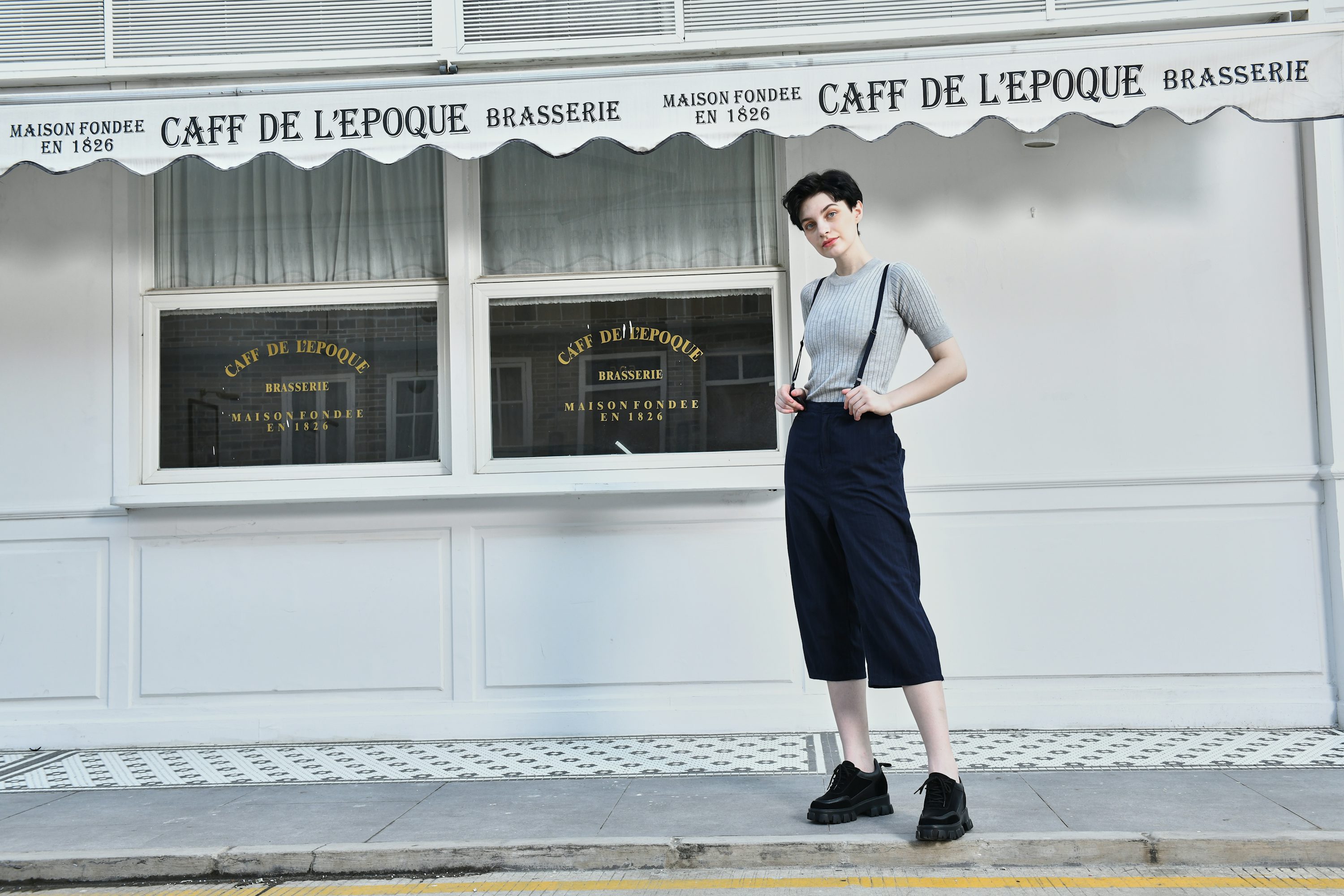 the image shows a young woman standing in front of a vintagestyle cafe. she's wearing a light grey ribbed sweater, dark navy cropped trousers held up by suspenders, and chunky black oxford shoes.  the cafe's facade is white with dark lettering in french, indicating the name "caff de l'epoque" and that it's a brasserie (a type of restaurant). the woman's style is reminiscent of 1920s or 1930s fashion.  the overall scene is aesthetically pleasing, with a contrast between the modern woman and the historical setting.
